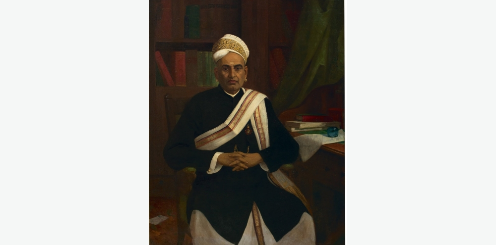 A traditionally dressed seated man with clasped hands faces us. He wears a white and gold headdress, gold earrings and a medal on his chest. Behind him is a desk with books and a sheet of paper.