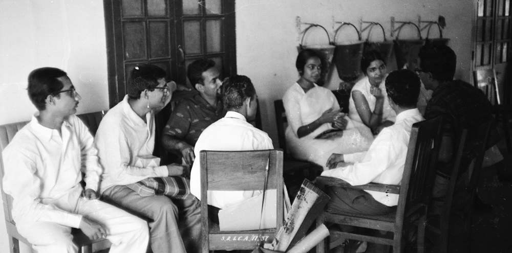A group of artists engage in conversation while sitting on chairs and benches facing each other in a corridor.