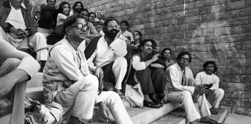 Multiple men and women sit on the steps of a staircase and gaze at something beyond the frame towards the right.