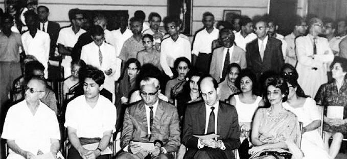 A large group of seated and standing people formally dressed as they look at something beyond the frame to our left.