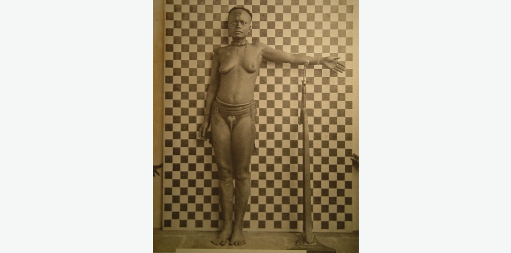 A nude woman with minimal jewellery stands in front of a chequered background with her outstretched left hand resting on top of a stand. The chequered background is held up by two arms from outside the frame.