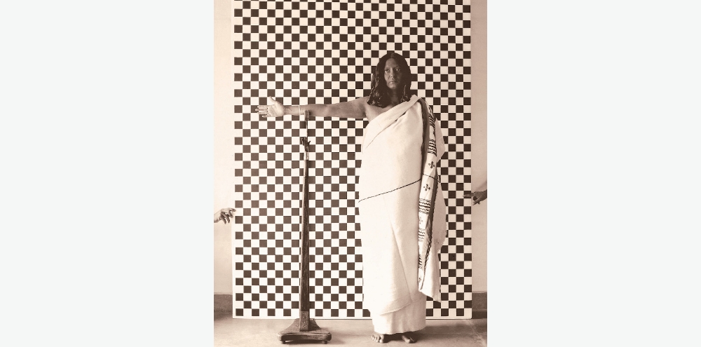 A woman in a white garment stands in front of a chequered background with her outstretched right hand resting on top of a stand. The chequered background is held up by two arms from outside the frame.