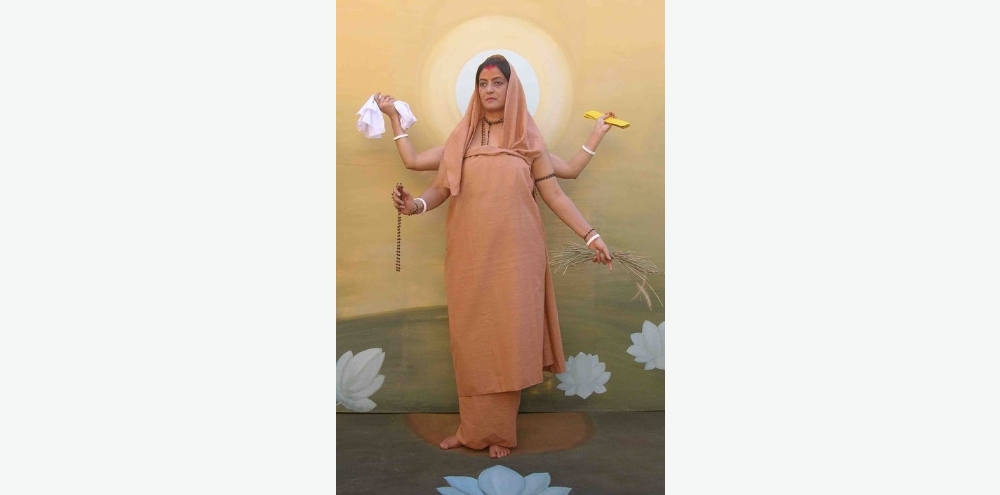 Photograph of a four-armed woman in a saffron garment standing in front of an orange background containing painted white lotuses.