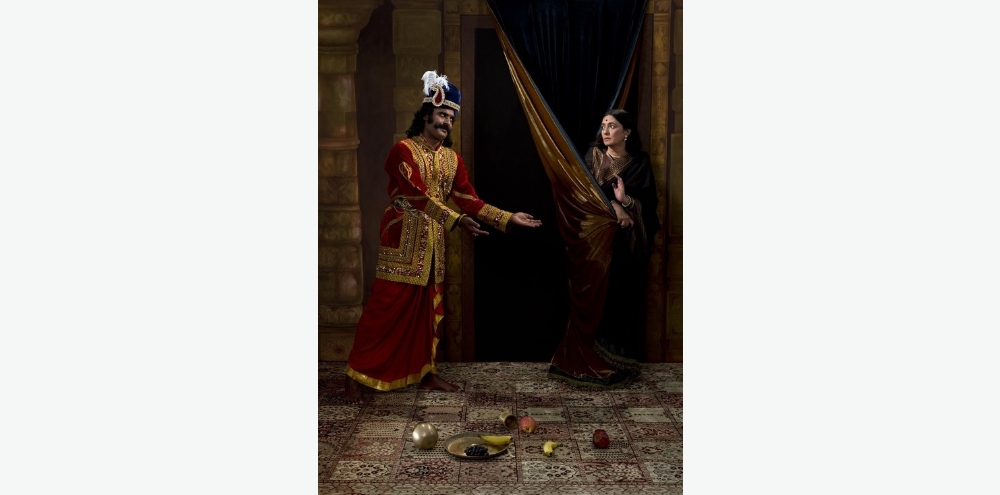 A photograph of a woman who hides behind a curtain and denies the advances of an ornately dressed man with a plate of fruits scattered in the foreground.