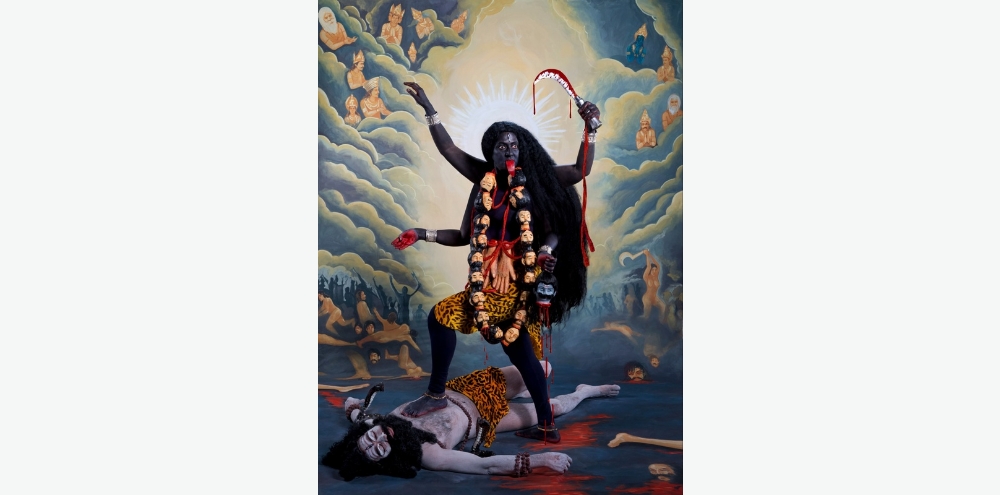 A photograph of a four-armed, dark blue-skinned woman wearing a skull garland and carrying multiple weapons points her bright red tongue out while stepping on a bearded man wearing tiger skin sprawled on the floor. Behind her, the painted background depicts a battle scene with gods as witnesses.