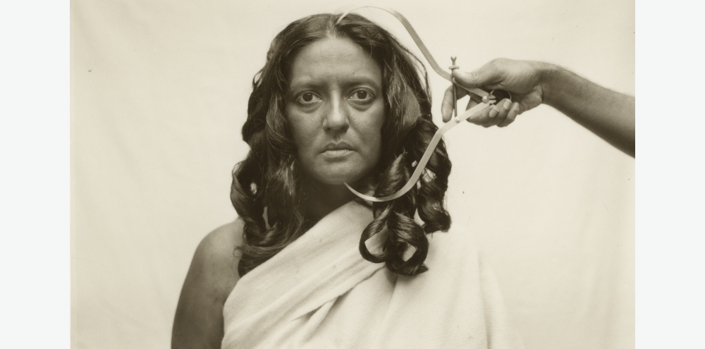 A headshot of a woman gazing intently at us while a hand on the right measures the length of her face with a tool.