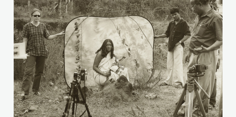 A seated woman, dressed in white, looks towards the camera amidst photography equipment and two people hold a screen in the back while a man stands on the right.