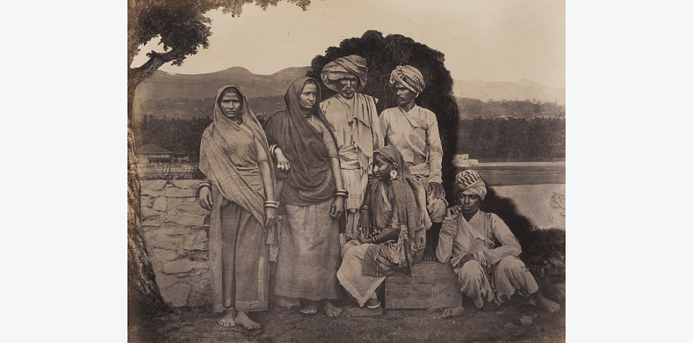 A group of three women in sarees and three men in kurtas, trousers and large turbans pose next to a tree.