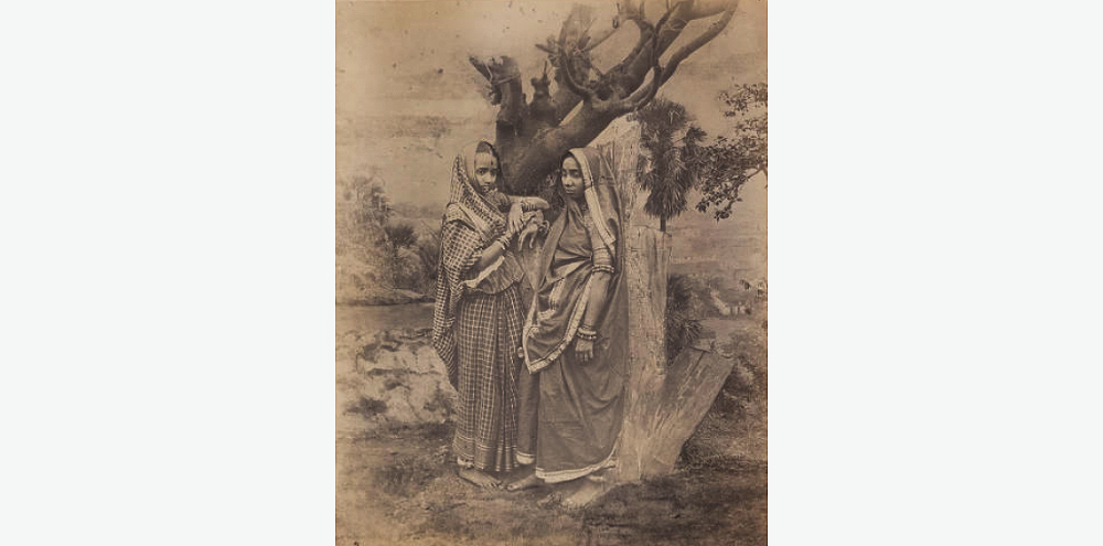 Two women in sarees and jewellery face each other while leaning against a tree.