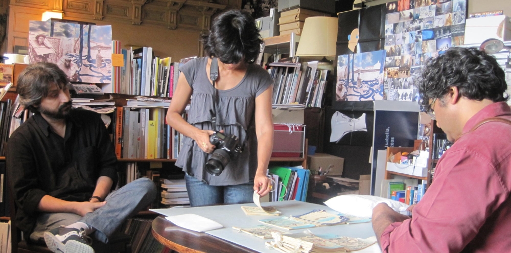 Two men and a woman surround a table with multiple booklets in a room full of drawings and books.