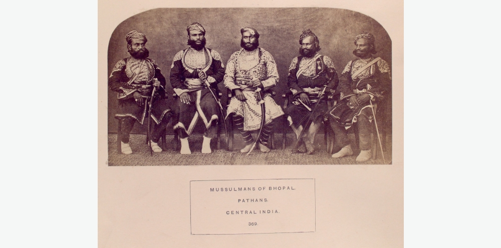 Five men with thick moustaches dressed in angarkhas and turbans, armed with swords sit next to each other and pose for the camera.