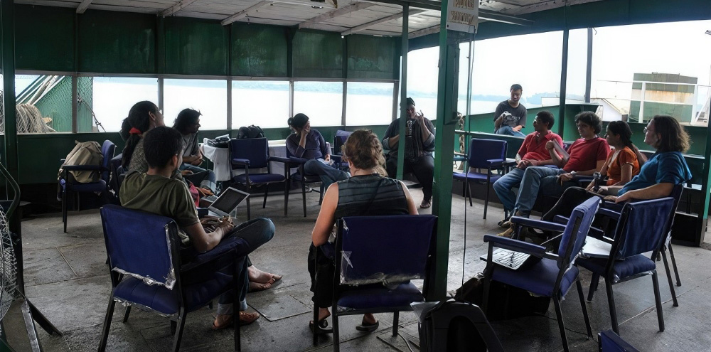 A group of people sitting in a circle face each other and listen to a man speak in a boat surrounded by water.