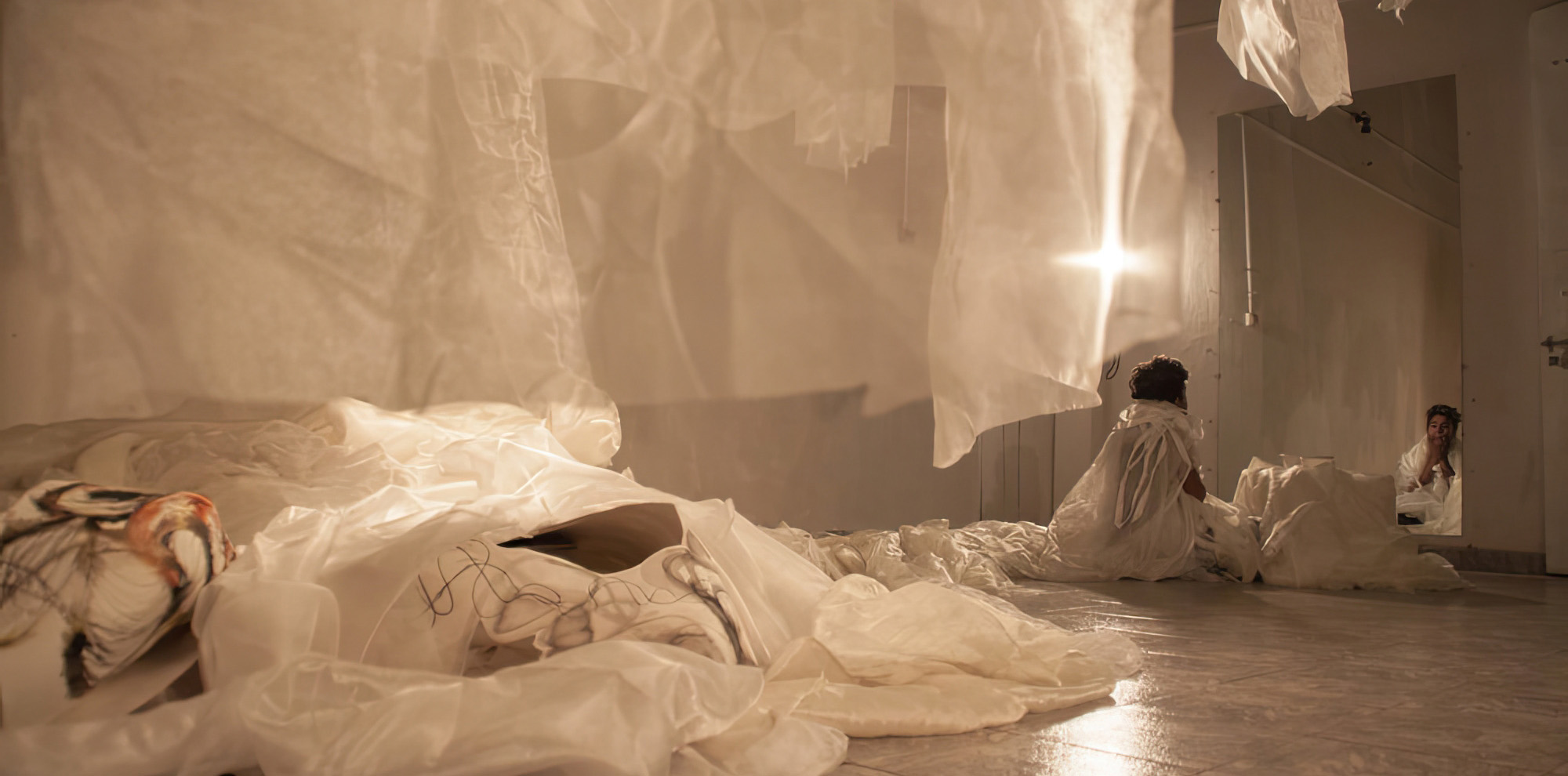 An artist looks at their reflection in the mirror on the right of the frame in a room that contains sheer white cloth hung from the ceiling.