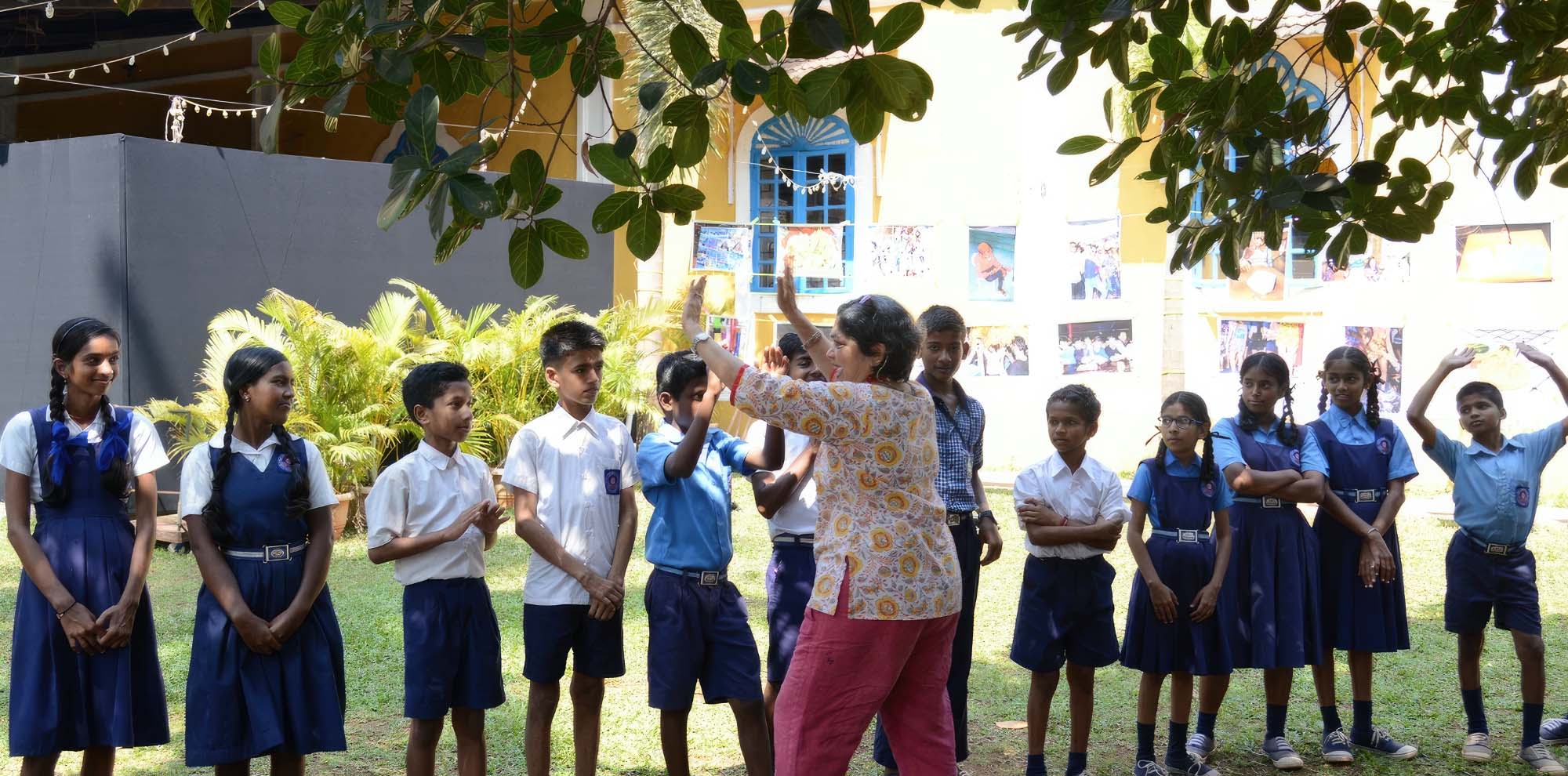 With hanging photographs and a yellow building in the background, a group of schoolchildren look at a woman holding her hands in the air.