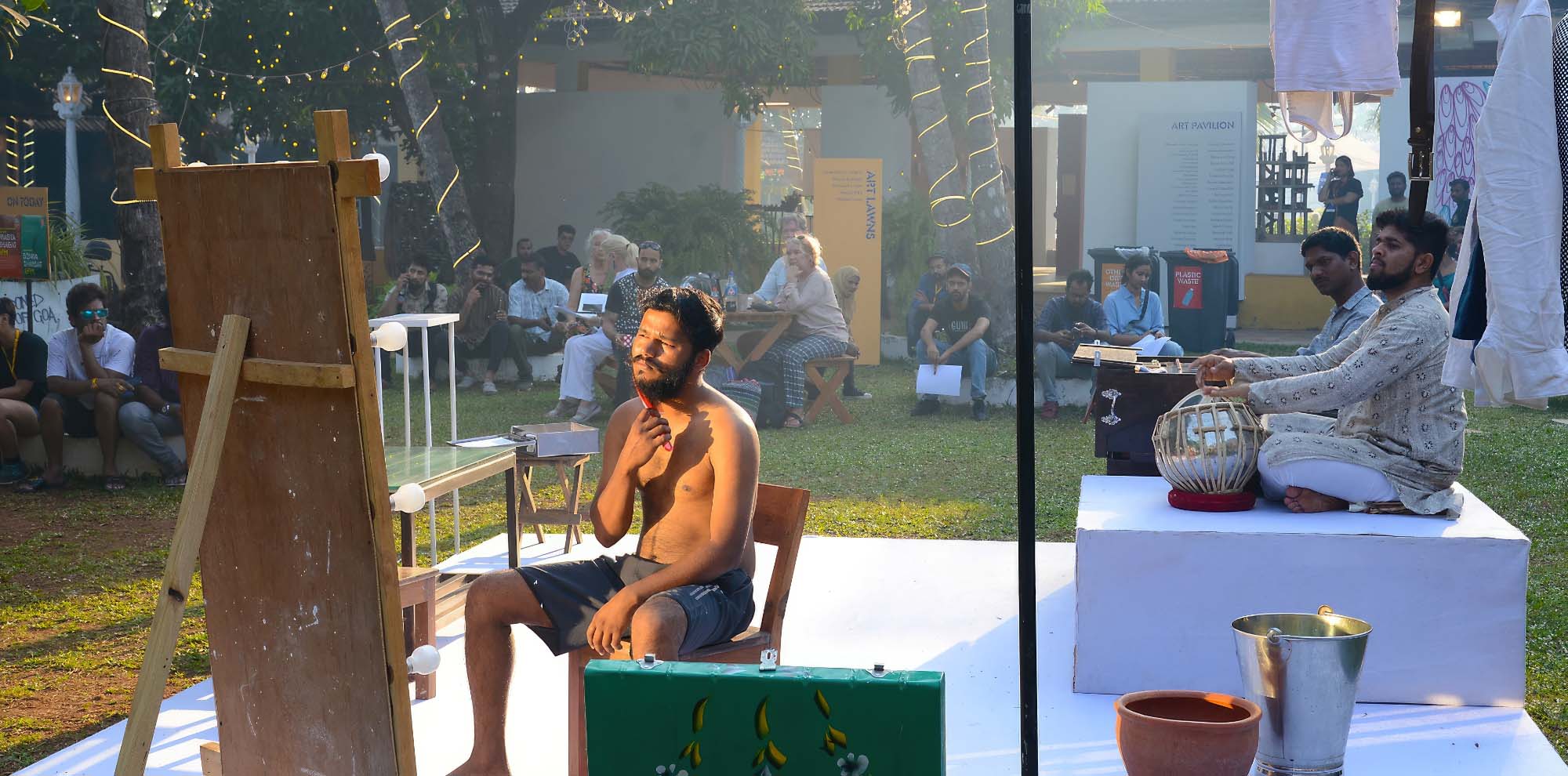 A man shaves his beard on the stage while two musicians are playing and an audience is overlooking the scene in the background.