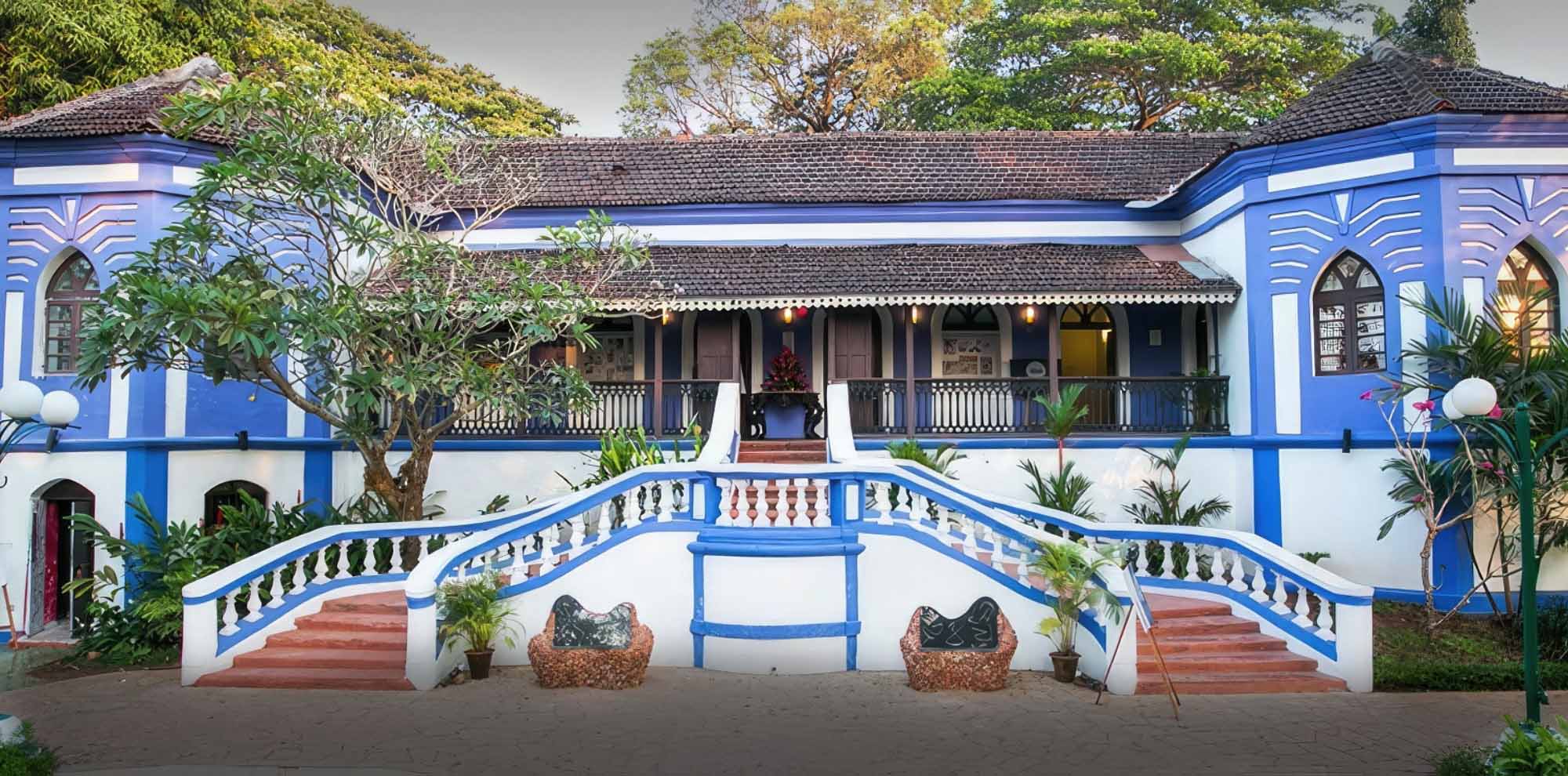The front facade of a large white and blue curved building features a double staircase surrounded by trees.