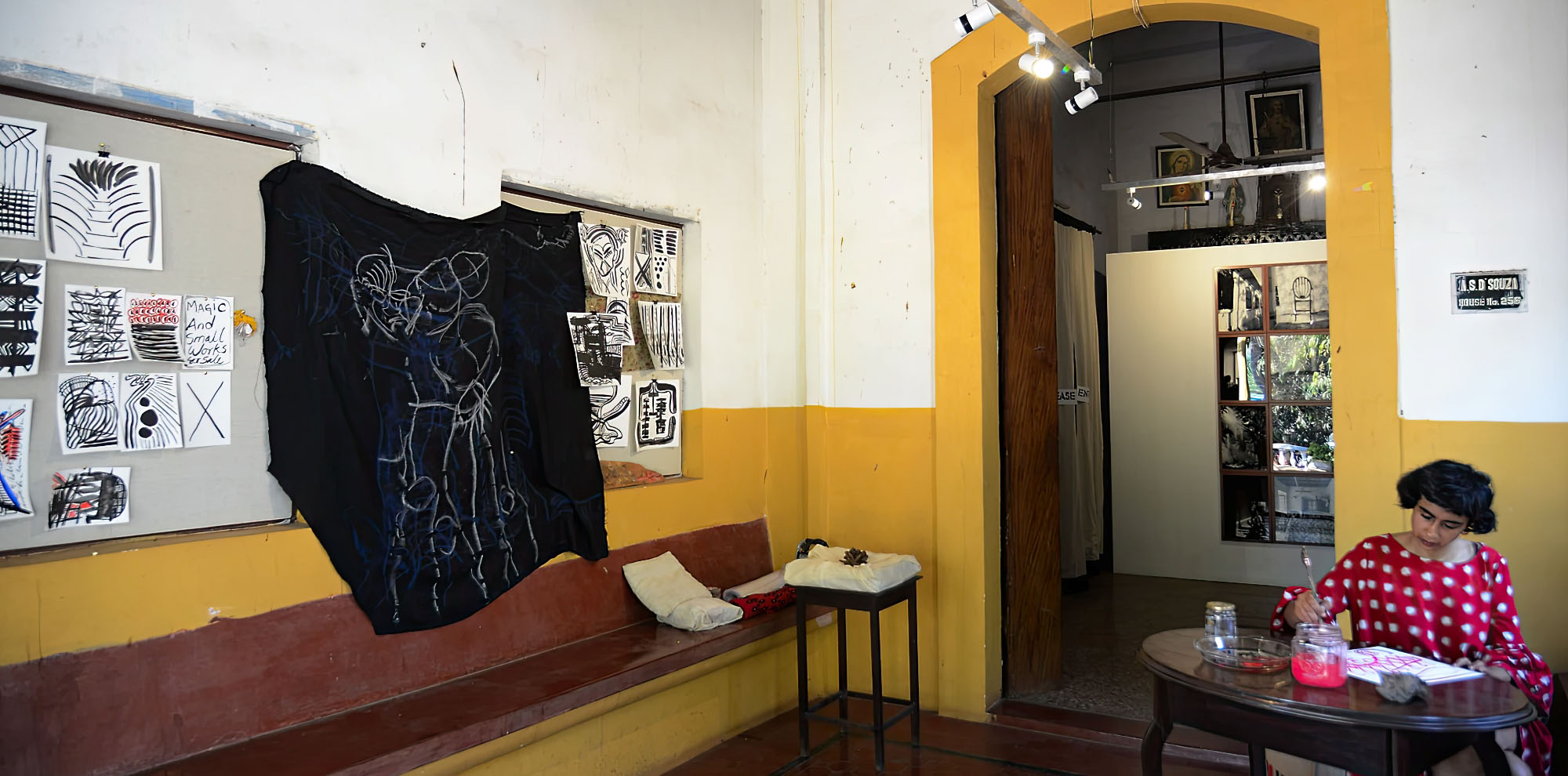 A woman sits and paints at a desk on the right side of the room that also contains several drawings on paper and a black cloth hanging from the wall.
