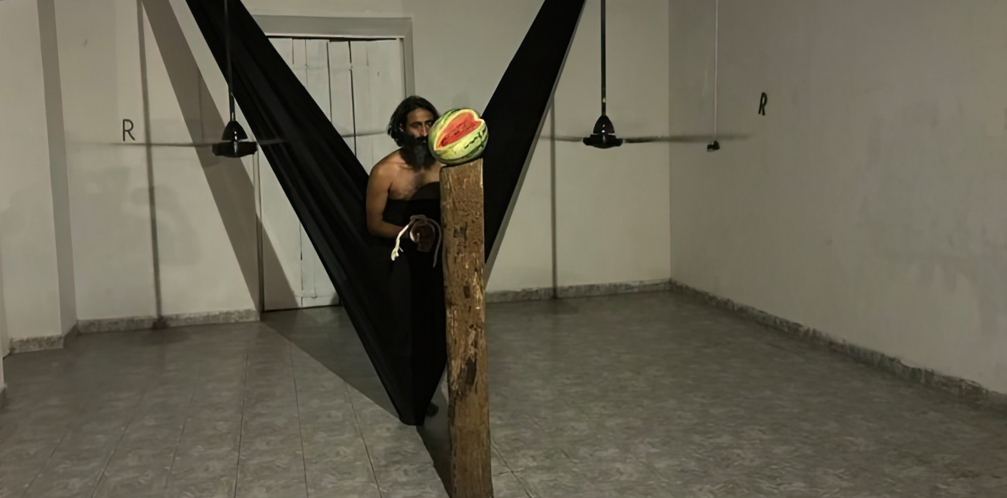 A figure clad in black garments stands behind two low hanging fans. A sliced watermelon is kept on a wooden pedestal in the foreground.