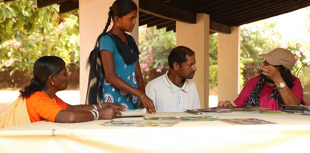 A group of three women and a man sit around a table scattered with artwork on a verandah.