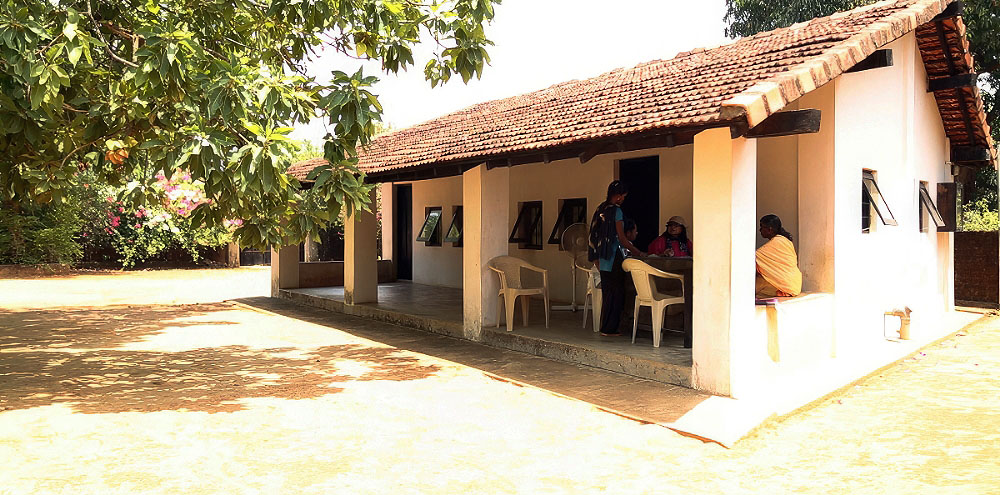 A group of people engage in conversation on the verandah of a tiled roof building surrounded by trees.