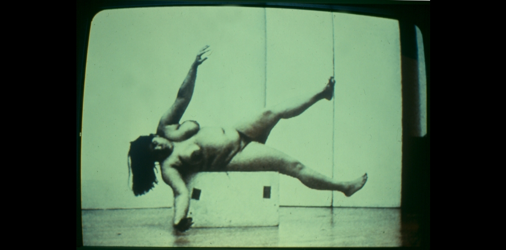 A nude woman lying down and attempting to balance herself on a stool.