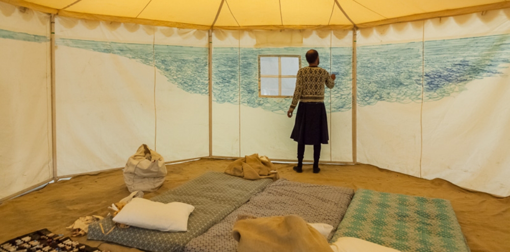 A man draws dense abstract blue lines on the interior of a tent with three futons and pillows in the foreground.