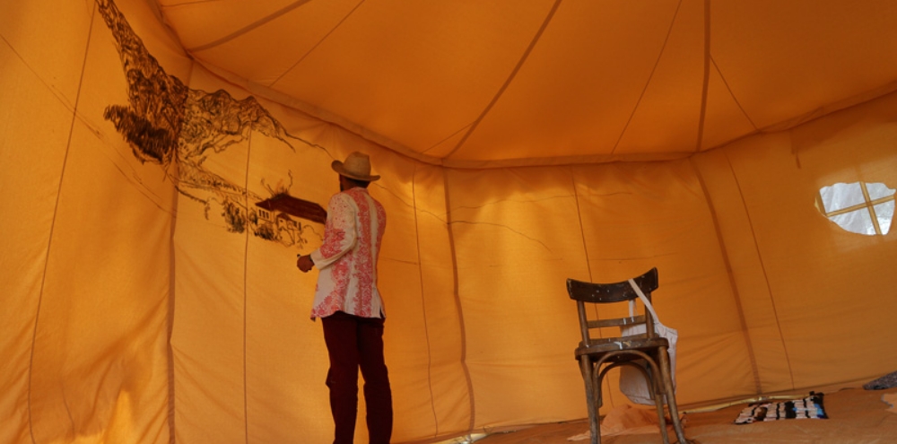 A man paints a landscape with mountains, trees and a house on the interior of a tent.