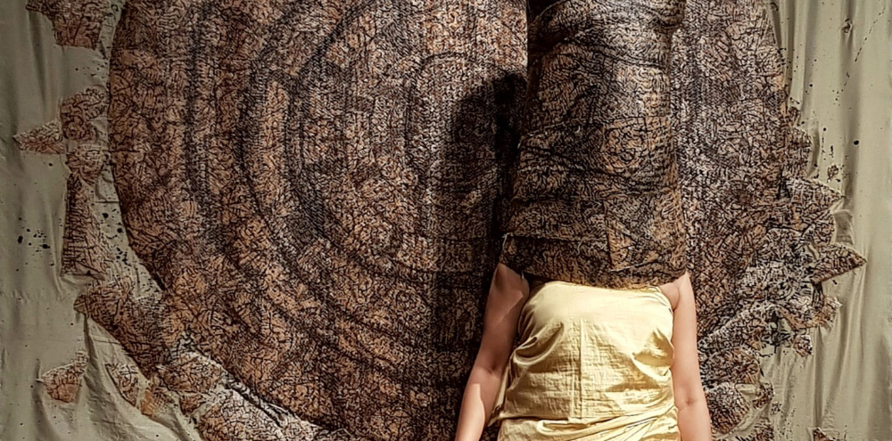 A woman in a yellow garment has her face wrapped in a painting that continues on the wall behind her.