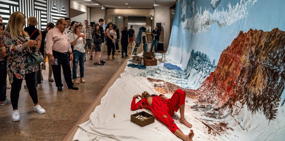 A man lies down in front of a large painted landscape with art supplies around him while an audience observes him from the left.