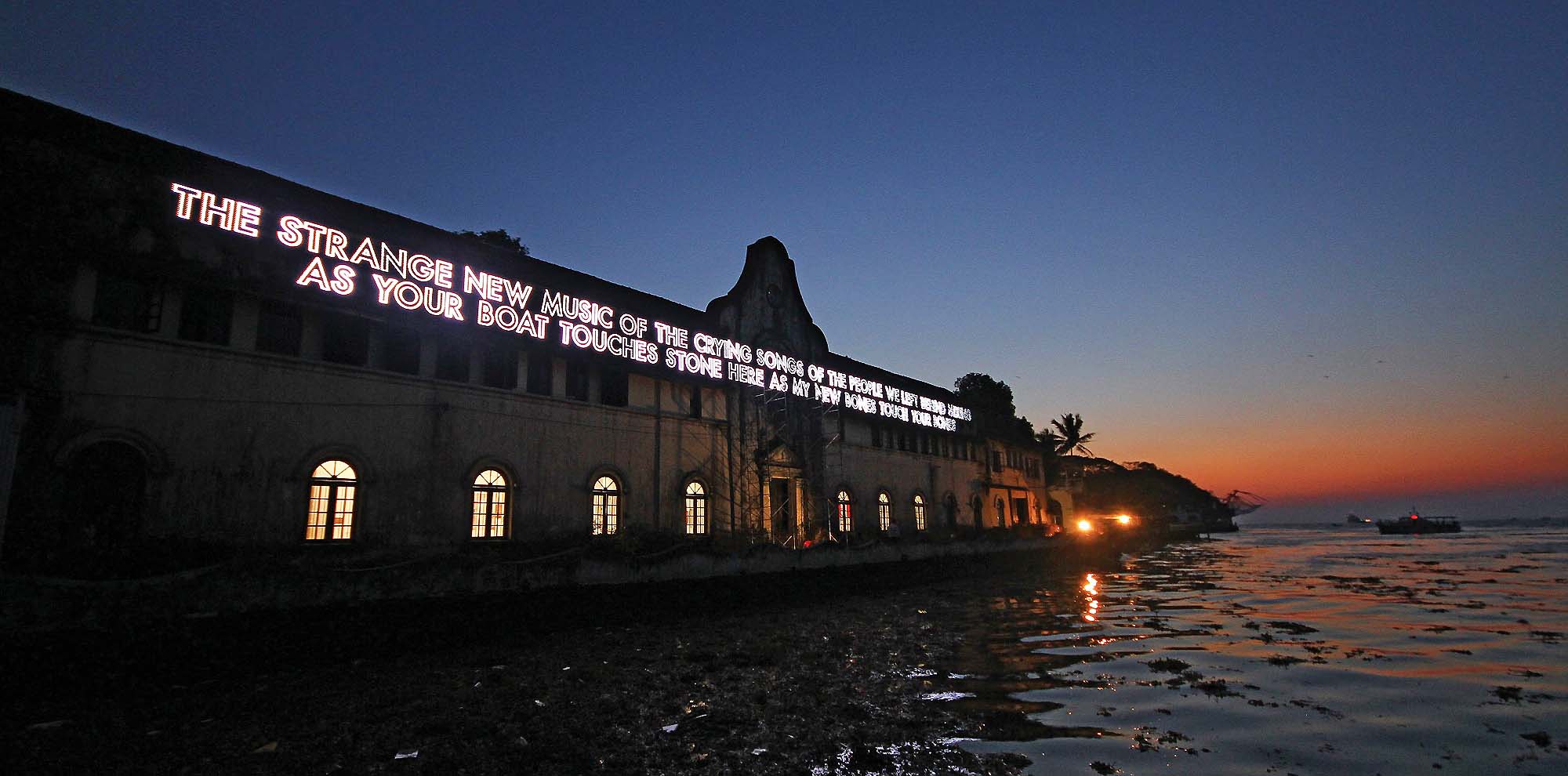 A light installation of text is displayed on the exterior of a building facing open waters.