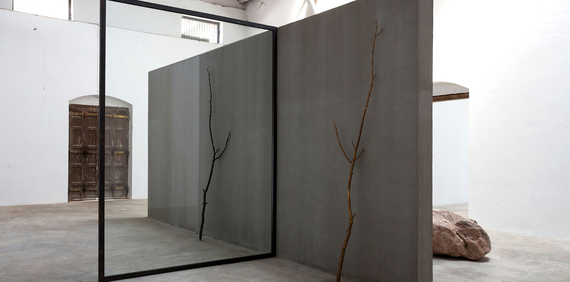 A branch and its exact replica leaning on either side of a concrete wall separated by a mirror.