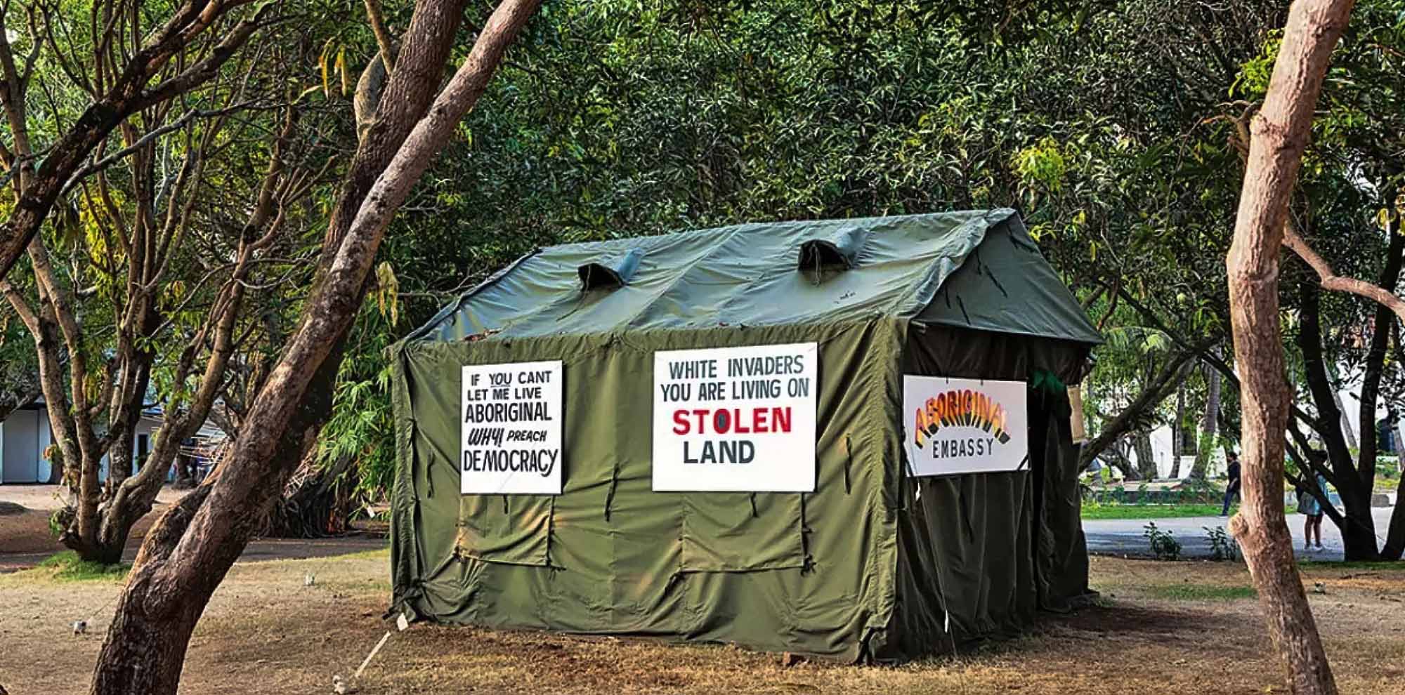 A green tent with various anti-colonial signs stands in the midst of trees.