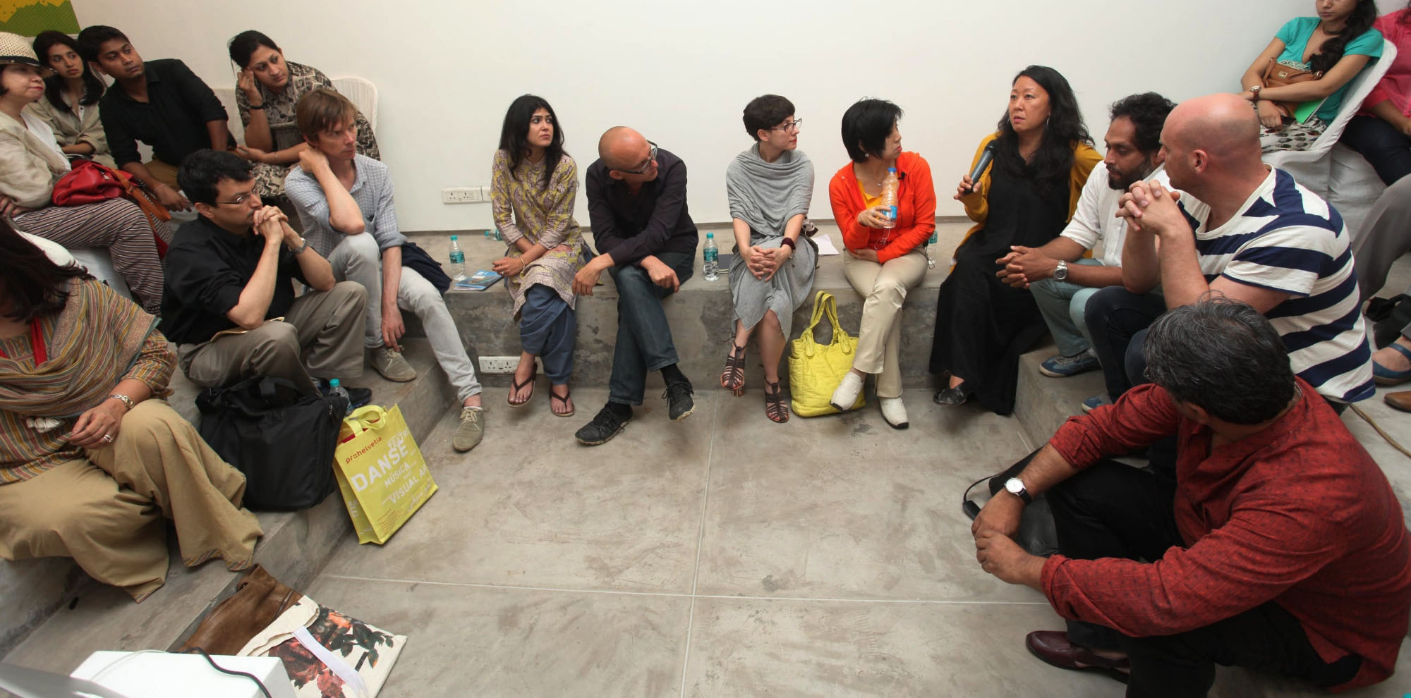 A large group of seated people listen to a woman talk with a microphone.