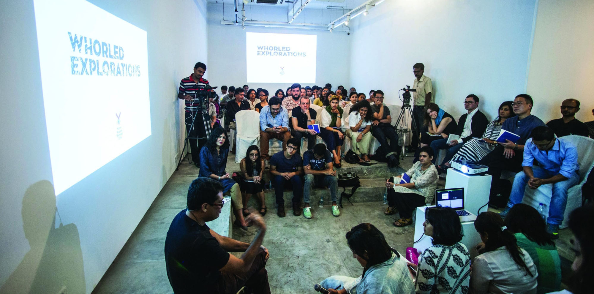 A large group of seated people pay attention to a man in front of a video projection on a wall.