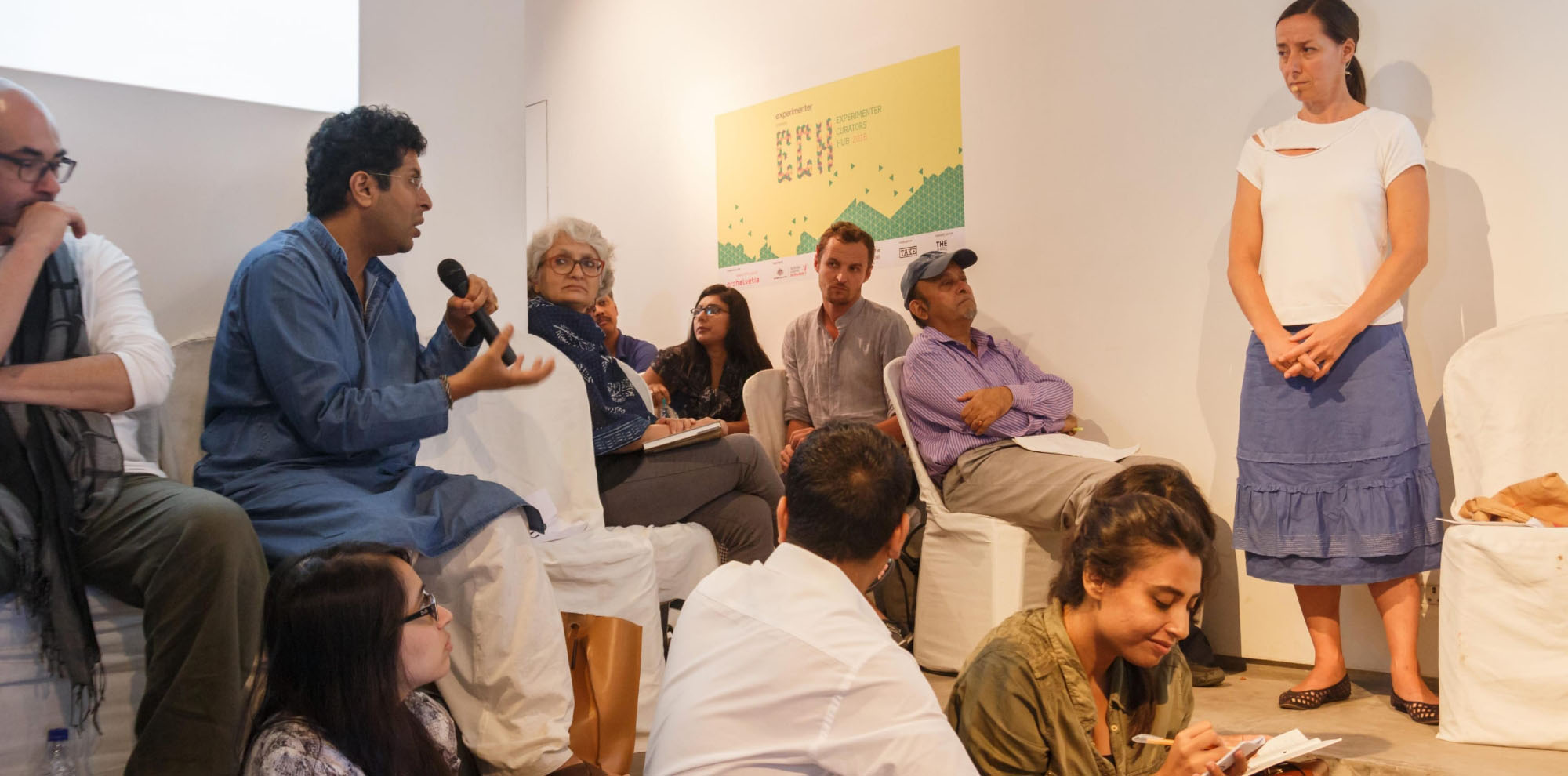 A man engages in conversation with a standing woman in the middle of a crowded room filled with seated people.