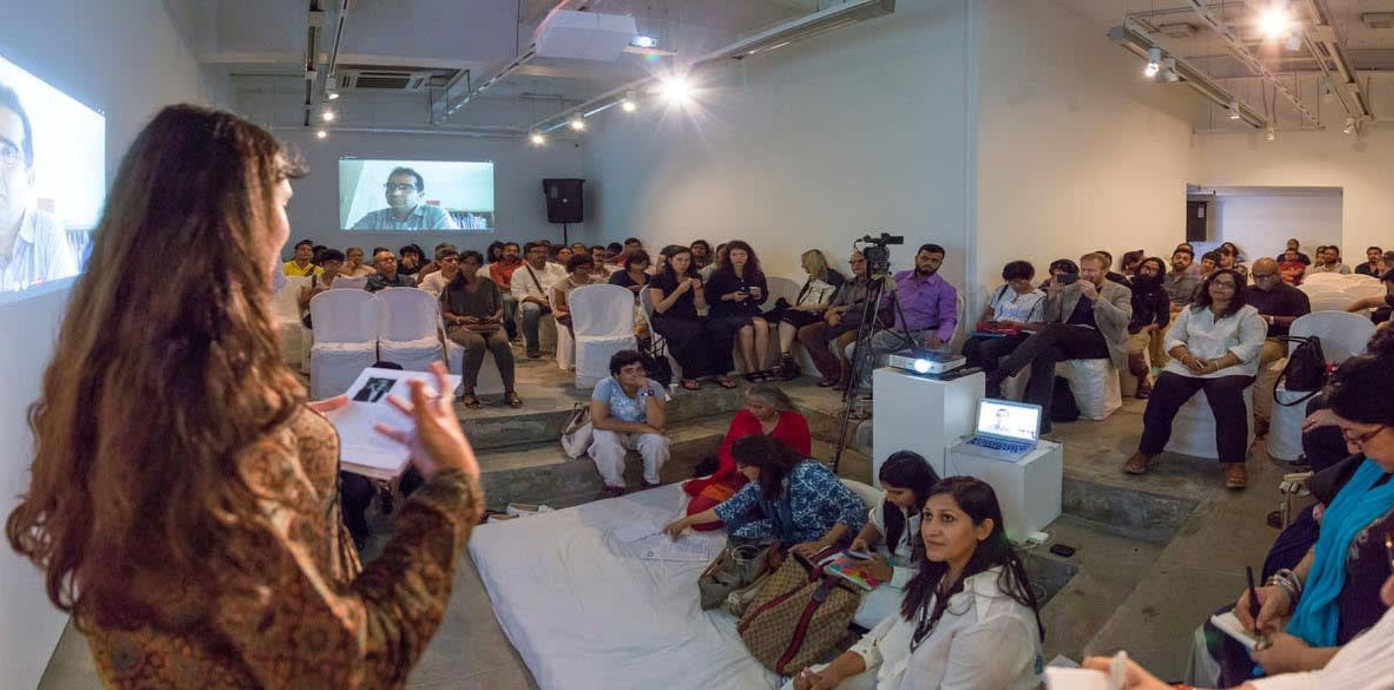A woman presents to a large group of seated people with a video projection of a man in the background.