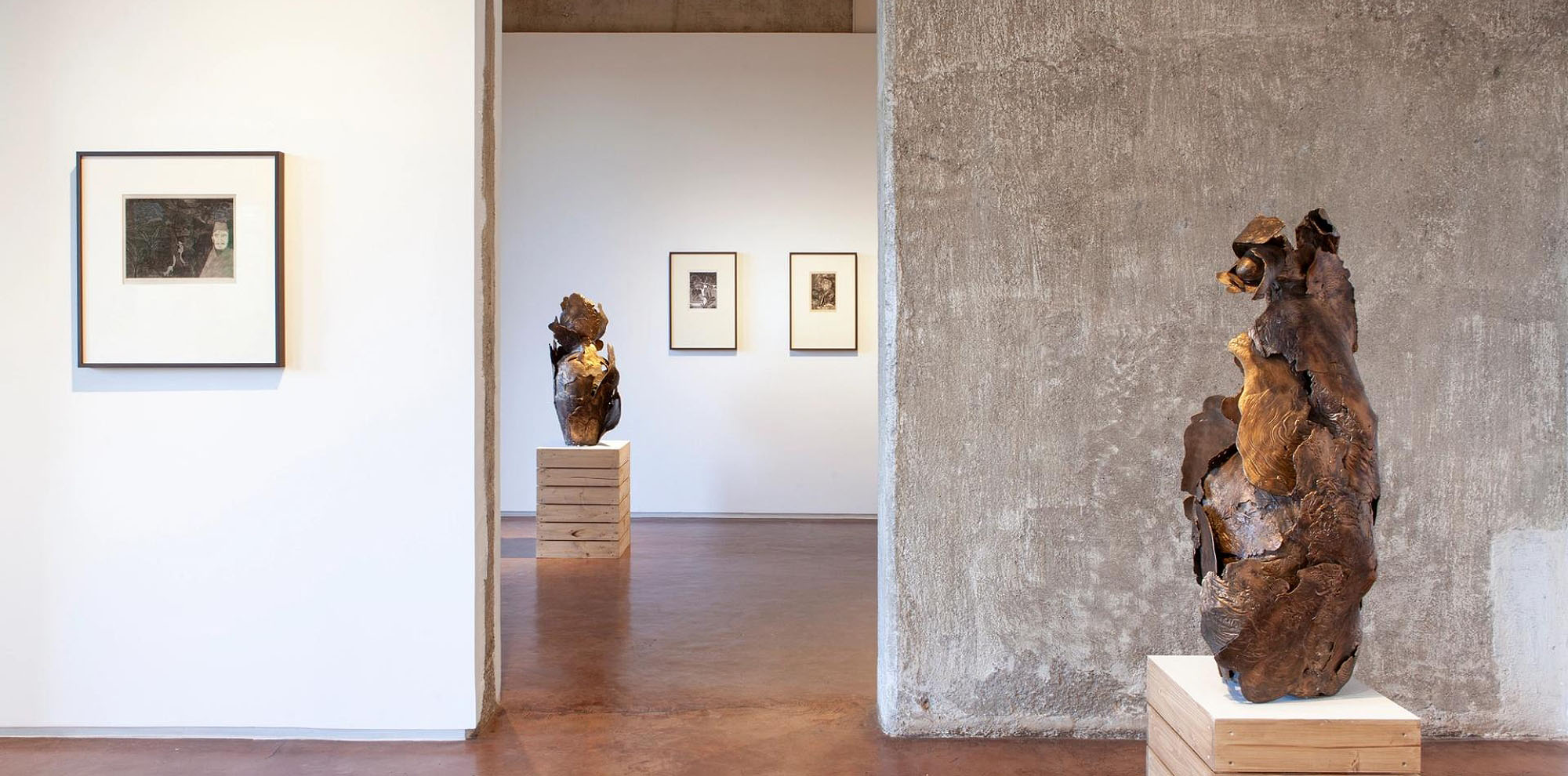 Several framed artworks hang on white walls surrounded by abstract metal sculptures on pedestals in an exhibition space.