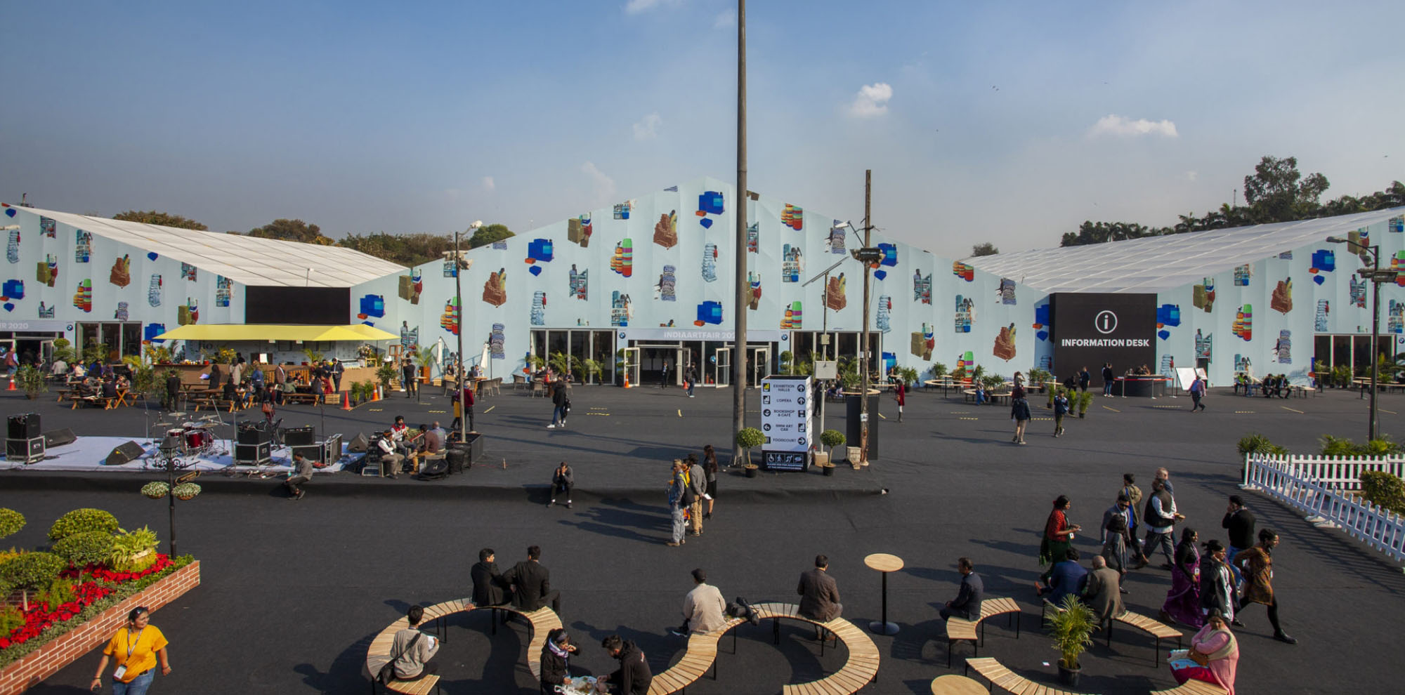The exterior of a large tent with groups of people walking or sitting on benches is found in the foreground of the image.