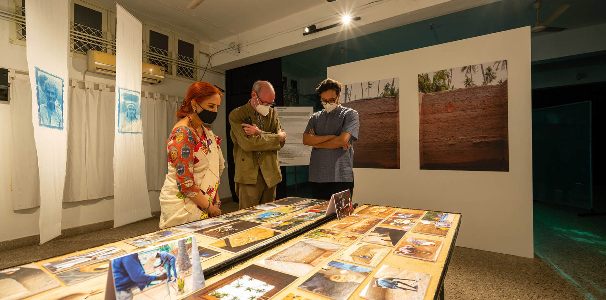 Two men and a woman observe a table full of photographs in an exhibition space.