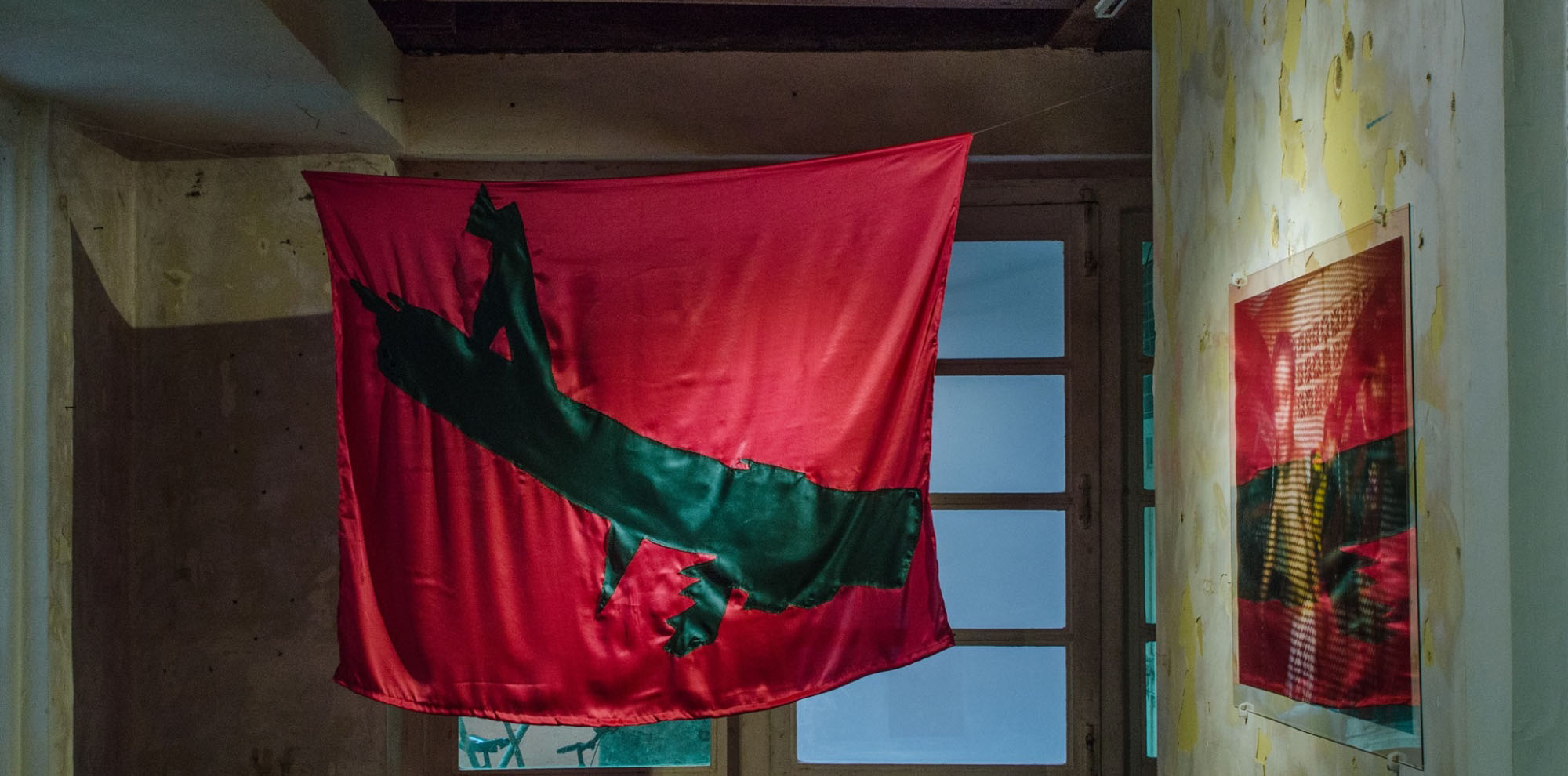 A red hanging cloth featuring green imagery whose pattern is mirrored on a photograph of a boy on our right within an exhibition space.
