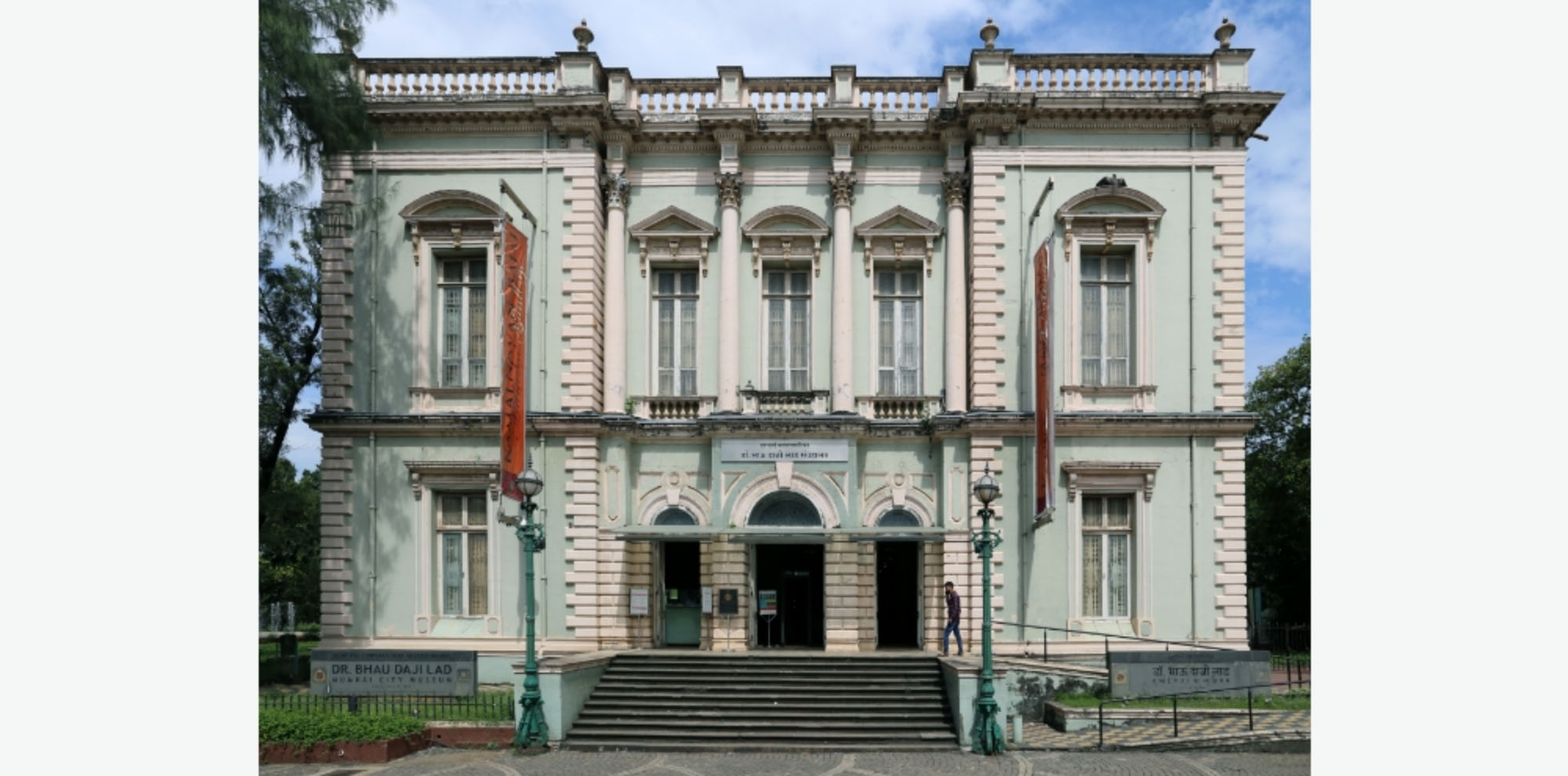 The front facade of a pale blue and white building and a sign with the English text, ‘Dr Bhau Daji Lad Mumbai City Museum’.