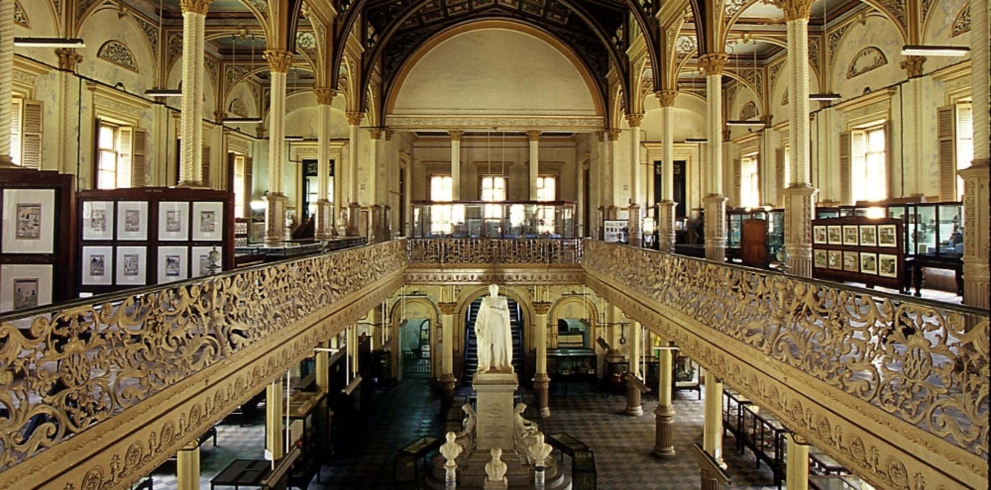 A long hall with yellow walls, pillars and railings and a large statue of a standing man in the centre.