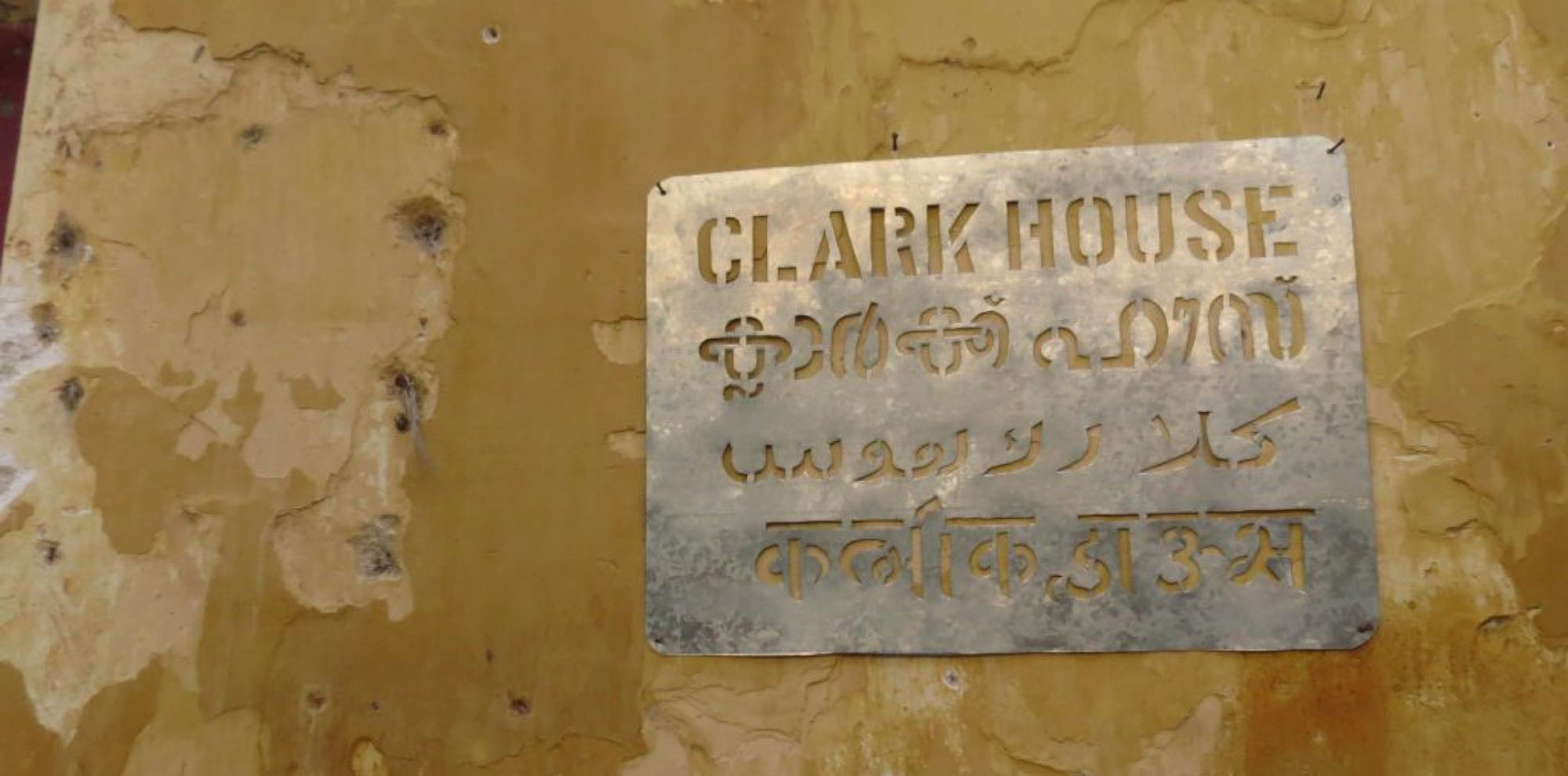 A metal signboard with the words. ‘Clark House’ is written in four languages on a yellow wall.