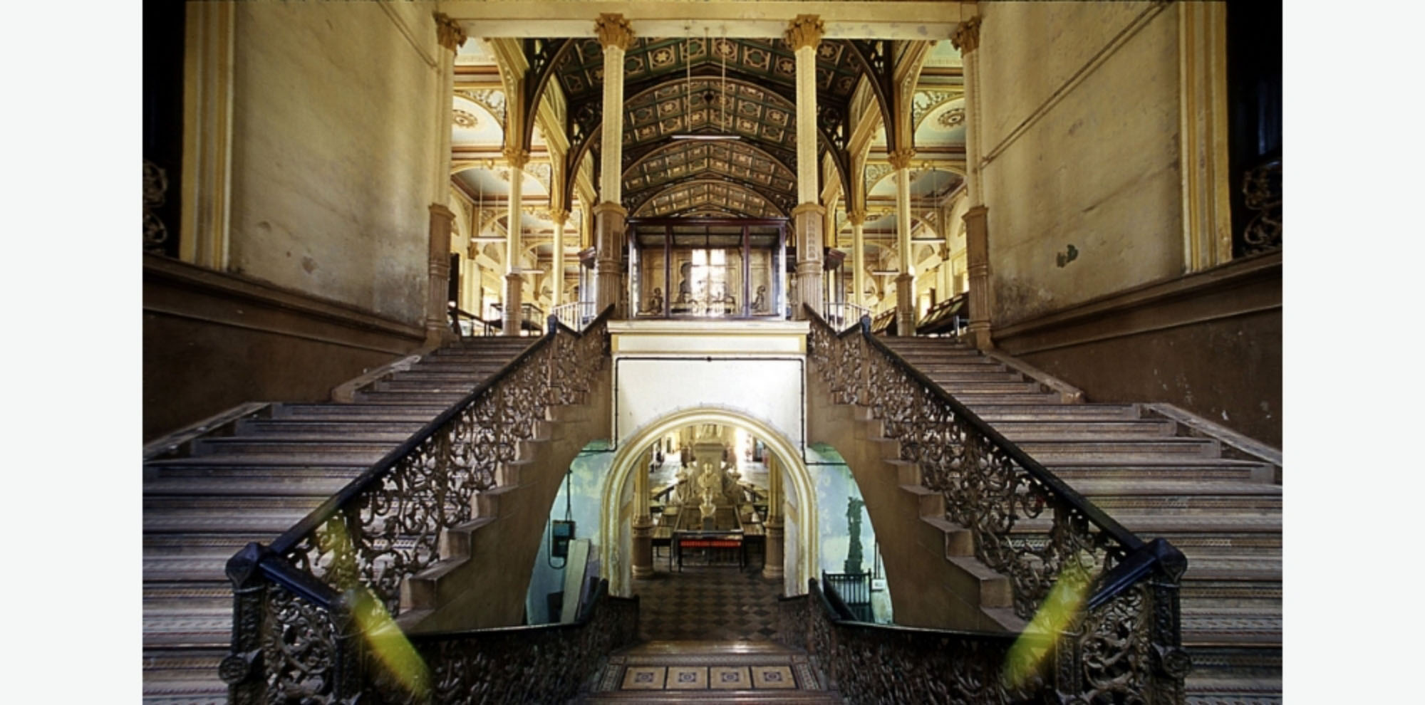 A dark stairwell with grey stairs, rusted railings and stained yellow walls with a view of the upper and lower exhibition halls.