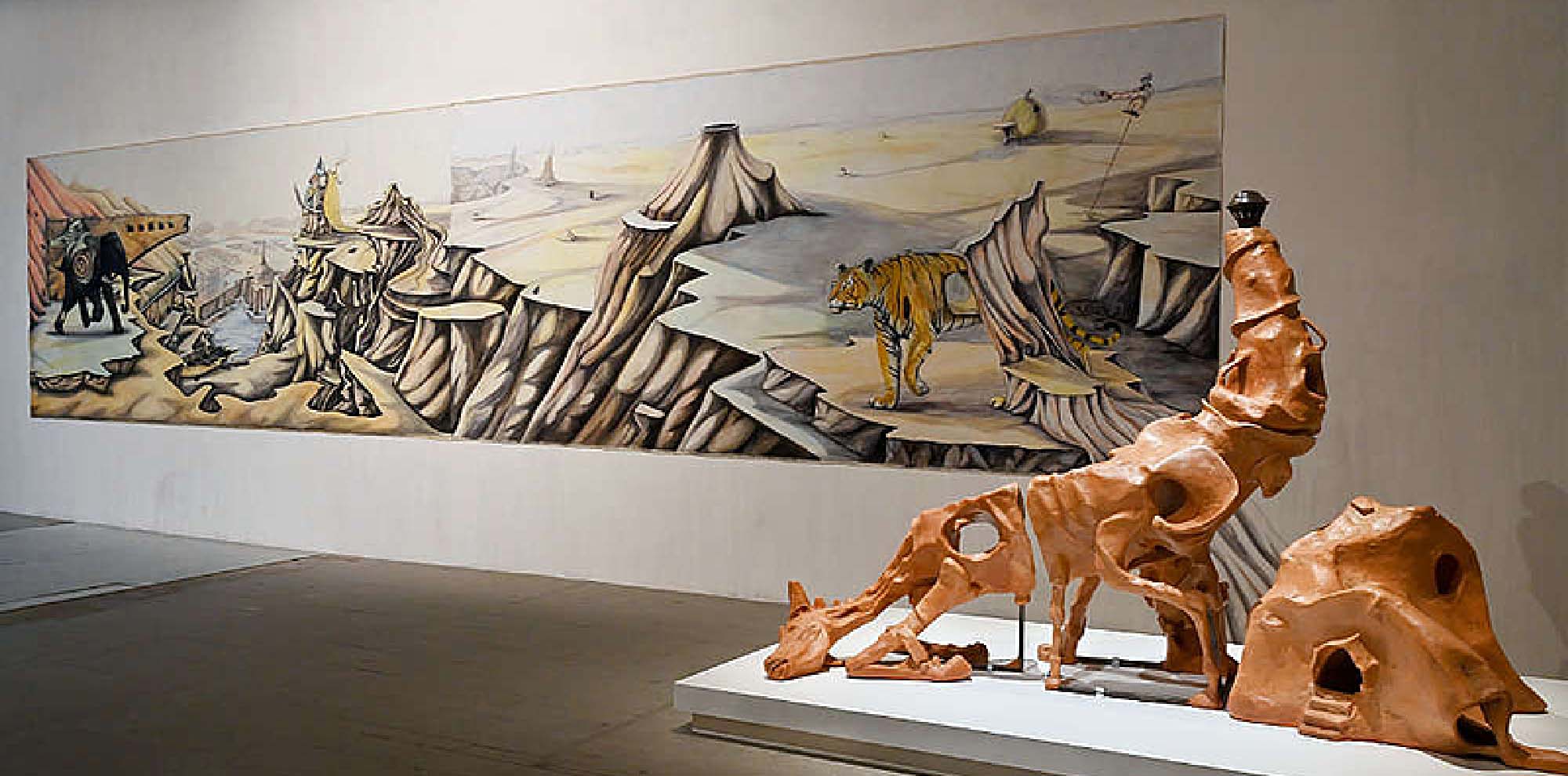 A large painted wall mural depicts the excavation of a ship in the desert. There is an elephant on the left and a tiger on the right with imaginary machines scattered about. A hybrid horse-scorpion sculpture is at the front of the painting next to a cave-like house.
