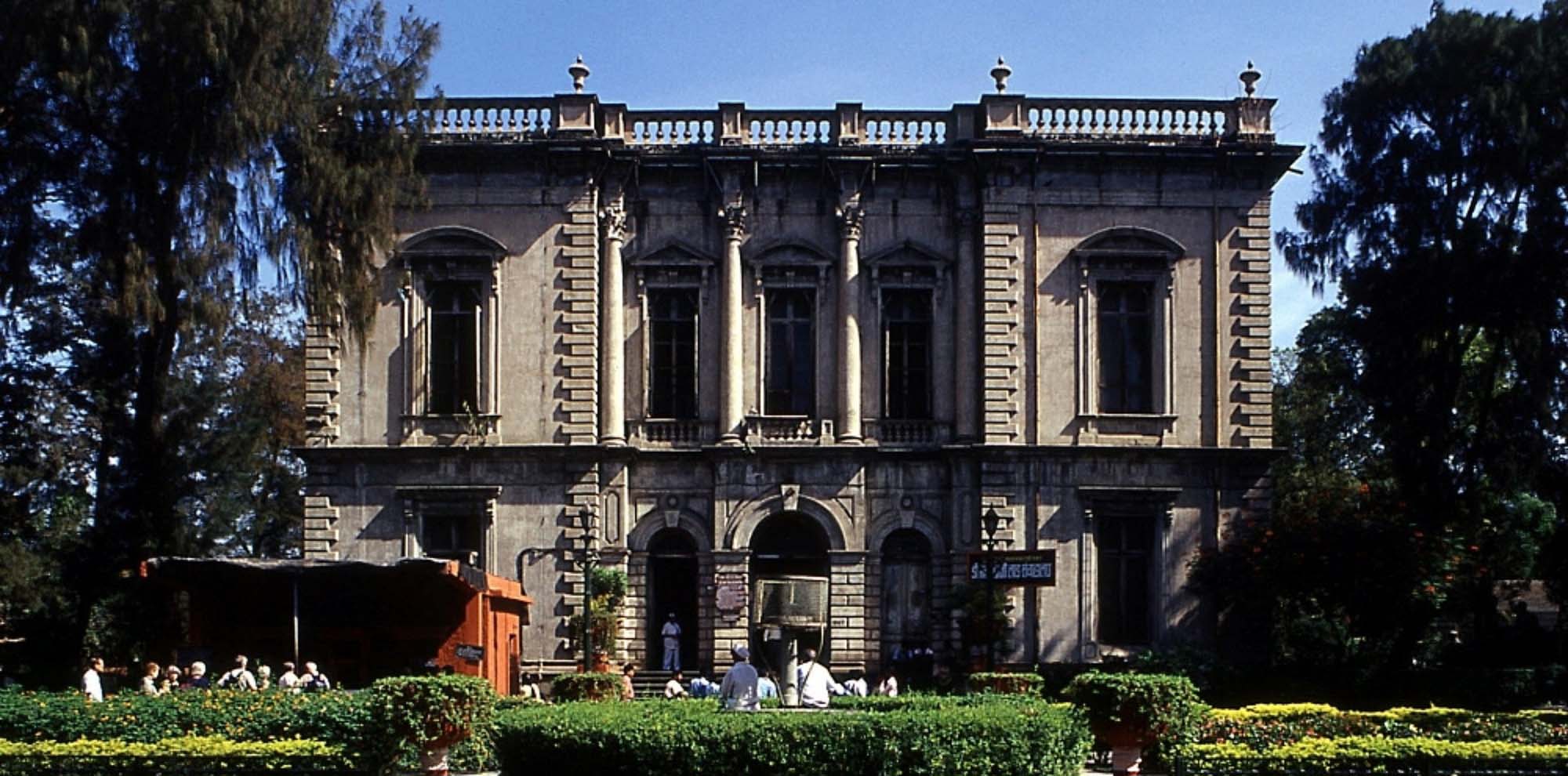 The front facade of an unpainted concrete building is surrounded by trees.