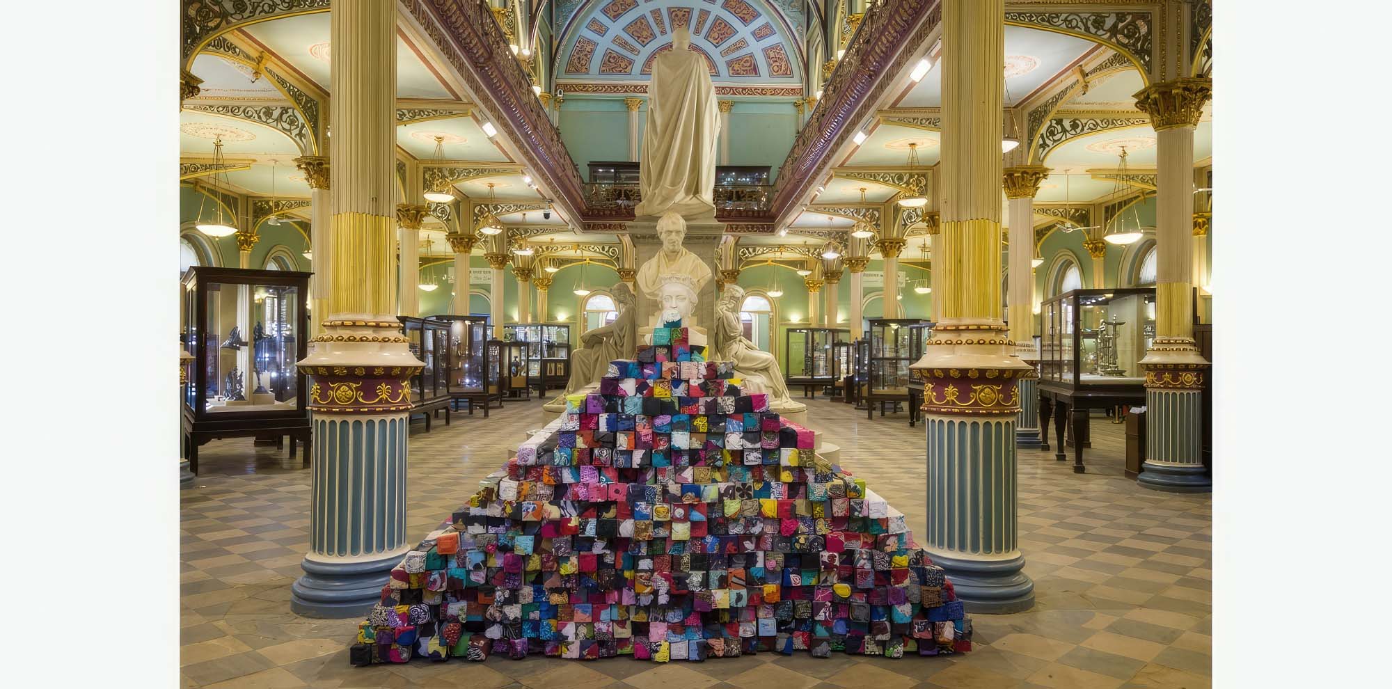 A pyramidal stack of blocks of compressed used cloth in a large open exhibition hall with a marble statue of a man.