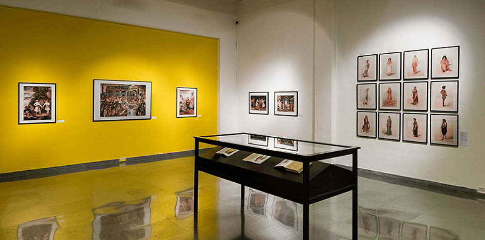 In the corner of the room, there is framed artwork on a white and yellow wall with a glass cabinet in the middle.