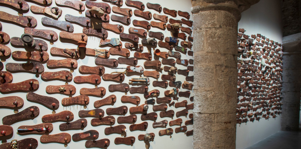Rows of toe-grip wooden sandals known as Khadau are displayed on a white wall with a stone pillar in front of it.