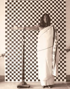 A woman in a white garment stands in front of a chequered background with her outstretched right hand resting on top of a stand. The chequered background is held up by two arms from outside the frame.
